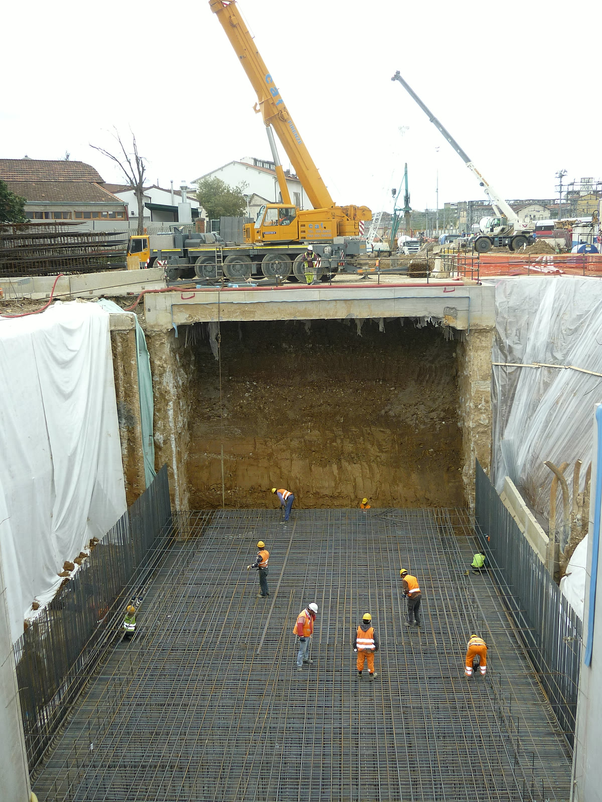 Deep excavation with workers placing the reinforcement of the foundation raft, cranes at ground level